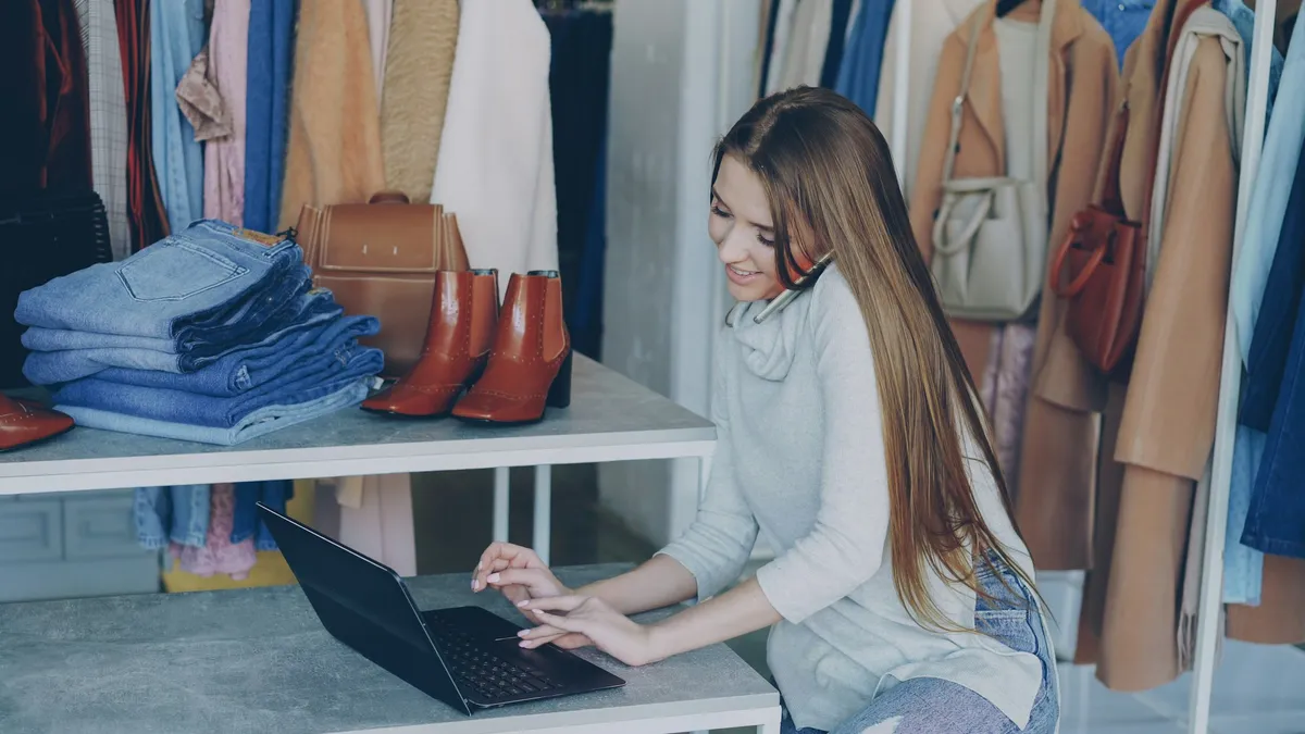 Jeune commerçante souriante qui travaille à son ordinateur portable, entourée des vêtements de sa boutique